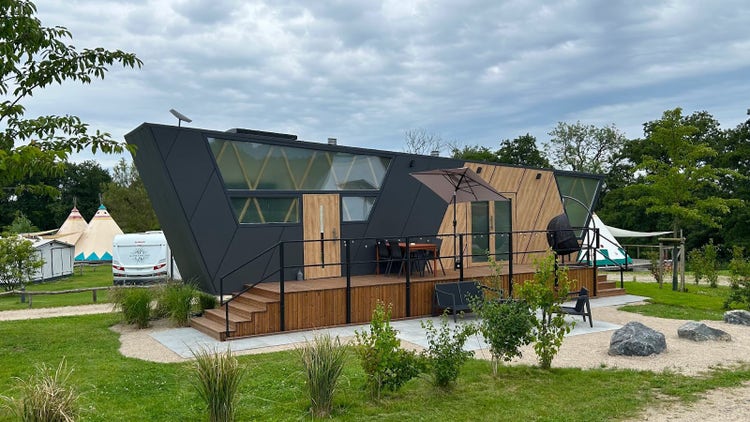 Modernes Tiny House mit ikonischer Form mit Holz- sowie Grauoptik und großzügiger Terrasse sowie Gartenmöbeln davor. Dieses ist platziert auf einem attraktiven Campingplatz mit Bäumen, einem See und blauem Himmel.