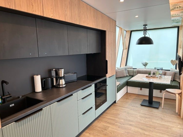 Modern kitchen and dining area with black countertops, sleek cabinets, and a cozy seating area around a wooden table. The space is illuminated by natural light from large windows.