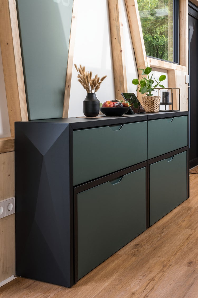 A modern wooden cabinet with green drawers, decorated with a plant, vase, and a bowl of fruit, is set against a wall in a bright, window-lit room.
