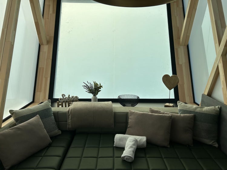 Modern tiny house with iconic shape and wooden as well as grey optic having a specious terrace in front of it including outdoor furniture. Placed in an appealing campsite with trees, a lake and blue sky.